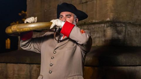 A man in a tricorn hat wearing a long brown coat with red cuffs and white gloves, blowing a curved horn.