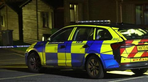 Police car parked on road with wooden lodge-style buildings along the far side and police tape across the road.