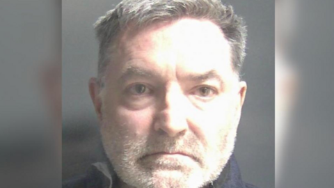 A man faces the camera in a mugshot. He has dark hair cut short at the sides, short white facial hair and he is wearing a black shirt