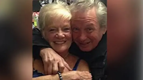 Sheila Jackson with blonde hair smiles as Eric Greener with grey hair has his arm around her neck in front of a bar. Eric is also smiling.