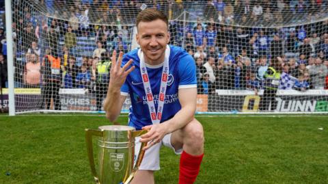 Lee Evans, in a Portsmouth kit, celebrating with the League One trophy which he has his left hand on while kneeling down on his right knee and holding up four fingers with his right hand
