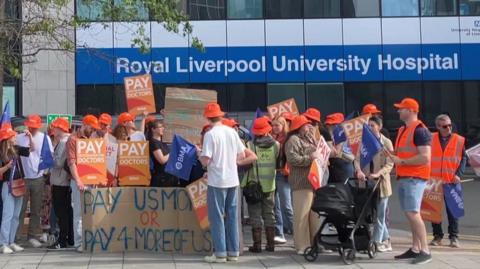 Row of people on strike wearing orange hats and waving various placards and signs outside the main entrance to Royal Liverpool University Hospital