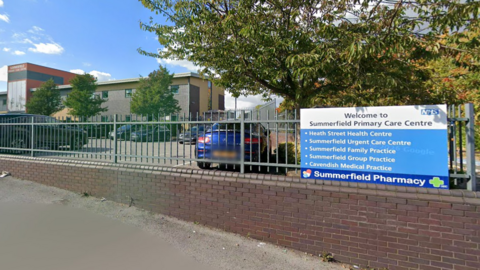 The front of Summerfield Primary Care Centre. There is a blue NHS sign with bullet points on and a sign for the pharmacy. It is on a grey fence and above a brick wall. Behind is a car park and a building. 