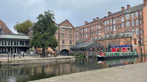 Ground-level view across Nottingham canal towards old multi-storey brick industrial buildings repurposed as bars, shops and accommodation.