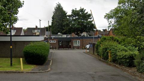 Exterior view of Wednesfield Mortuary, Alfred Squire Road, Wednesfield, Wolverhampton. It has brown front doors and surrounded by bushes and trees. there is a black and yellow striped barrier, which is up.