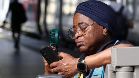 A railway worker uses an electric fan to cool herself down on a train platform in London.