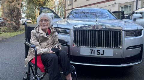 A woman with white hair and glasses in a light brown coat and black trousers, sitting in a wheeled chair in front of a large silver Rolls-Royce with the number plate 71 SJ