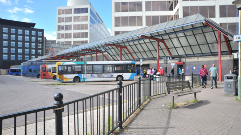 Image of buses lined up at Swindon Bus Station with people walking on the pavements. There are high-rise buildings in the background