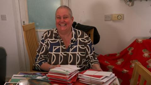 A woman with a buzzcut smiles at the camera. The table in front of her has loads of cards and letters. She has a green, blue and white patterned shirt on.