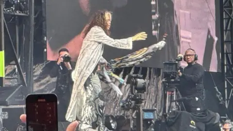 A man on stage with a photographer and camera operator behind him.