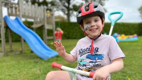 Samuel is wearing a red and black helmet and a light purple 'Racing Champion' t-shirt sits on a blue tricycle in a grassy backyard. The child has a colorful bandage on their right cheek and waves with their left hand. Behind them, playground equipment, another child in red, and scattered toys are visible.