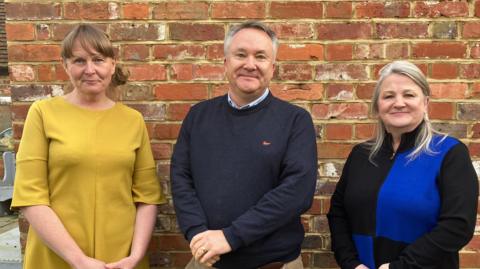 A picture of three people stood up against a wall of a pub. On the left, a Catherine Bunting has light brown hair tied back in a pony tail and is wearing a mustard yellow tunic. In the middle Stuart Wilson has greying hair and is wearing a dark blue jumper and light brown trousers. On the right, Anne-Marie Vladar has light blonde hair and is wearing a blue and black checked dress.