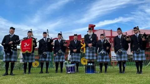 Nine people with bagpipes and drums, wearing Manx tartan kilts standing in a field. One has a Manx flag on his bagpipe.