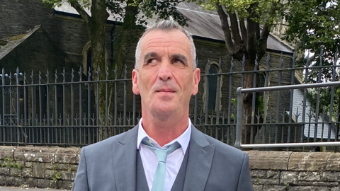 A blind man with spiked up white and grey hair. He is  wearing a grey two piece suite with a white collar shirt underneath a wait coast and a light sky blue slim tie. He is standing in front of a gothic looking church