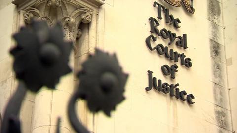 The stone facade of a building with "The Royal Courts of Justice" displayed in black lettering on the wall. There are floral iron decorations from the top of a gate in the foreground.