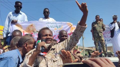 A man in military uniform in the foreground , holding a microphone, gestures. He is surrounded by Sudanese people attending a protest in support of the army in the eastern city of Gedaref on 22, February 2025