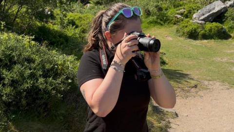 Jelly Boxall, with sunglasses on her head and her long, dark hair in a pony tail, is wearing a black t shirt and is taking a photo with her camera on Dartmoor. 