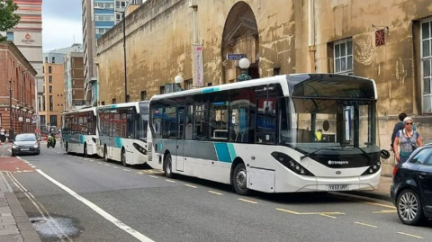 A line of buses parked on bus stops on a road in central Bristol. They are all single decker vehicles with white, blue and black liveries on them