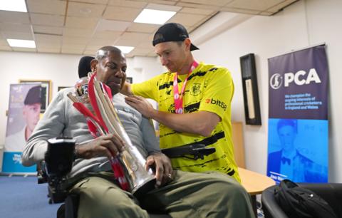 Syd Lawrence, in a wheelchair, holds the T20 Blast trophy after Gloucestershire won the 2024 final against Somerset. He is being presented with it by player James Bracey