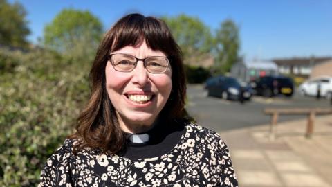 A dark-haired woman in a black and white flowery dress, with a clerical collar, smiling at the camera with bushes and parked cars behind her