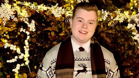teen/ harry with blonde hair wears a shirt and tie and brown scarf. His jumper has reindeer on. Background is gold and festive. This is his album cover.