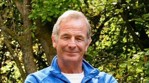 Robson Green. He is wearing a blue fleece and has grey hair. He is smiling at the camera.