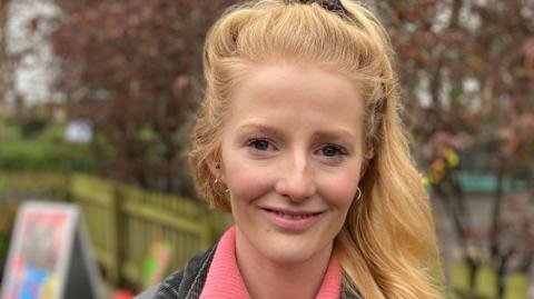 Hannah Spencer has long blonde hair and is wearing a black denim jacket and pink sweater. She is standing outdoors in a playground.