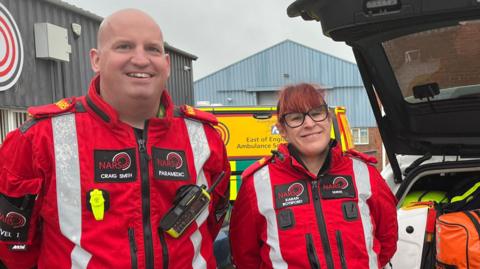 Craig and Karan are both wearing red jumpsuits with hi-vis strips. Craig is bald. Karan has red hair and is wearing black glasses.