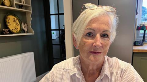 Julie Doorne sits in the kitchen at her Lincolnshire home. She has short, white hair and wears a white and grey striped blouse and pink-rimmed glasses on the top of her head.