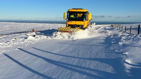 A yellow snowplough pushing through a thick blanket on snow on the Mountain Road. There are farmers' fenceposts at the side of the road.