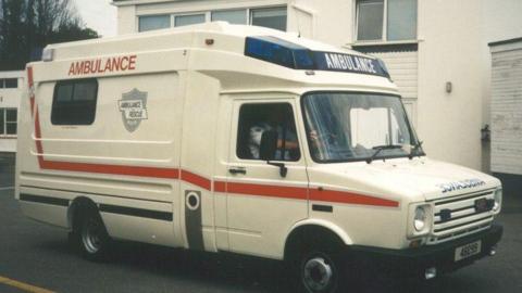 A white ambulance van with a red strip along it. It is an old picture. It is parked in front of a white building. 
