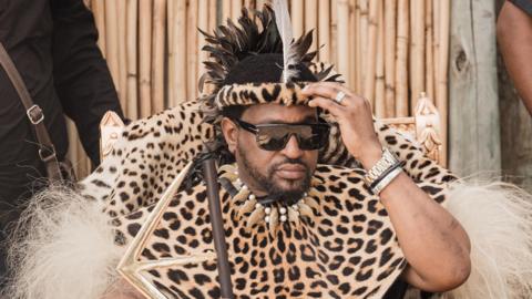 King Misizulu wearing traditional leopard-skin clothing and holding a stick. He is wearing dark glasses and is touching his head