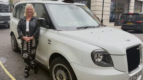 A blonde woman stands in front of a white car