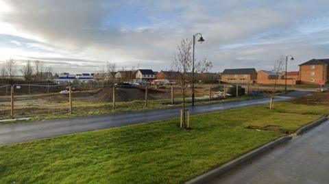 A building site where the proposed nursery and shop could be built. It has a large pile of mud in the middle, is fenced off and is next to a road that leads to houses.