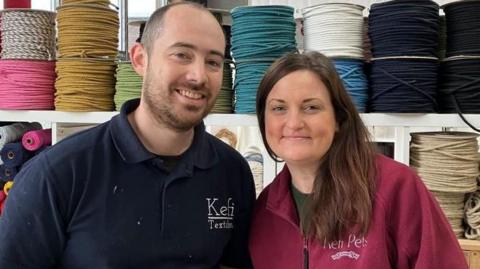 Andrew and Heather Cumpstone stand side by side, smiling, against a backdrop of colourful yarn on bobbins. Andrew wears a navy polo shirt with a white stitched logo reading Kefi Textiles on the chest. Heather has long brown hair and wears a maroon fleece white stitched logo reading Kefi Pets on the chest.