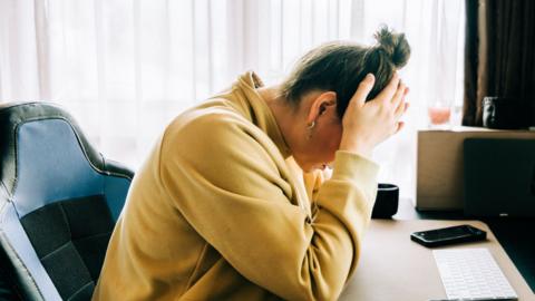 A woman in a yellow jumper sits at her desk with her head in her hands. 