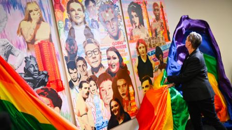 A large, brightly coloured artwork including figures from popular culture and landmarks from around the North East. A person is draping a rainbow Pride flag across the front of the artwork.