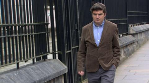 Michael McMonagle walking along the pavement beside a low stone wall with tall black metal railings. He is walking with one hand in his pocket, looking down. He is wearing a brown jacket, blue shirt, and grey suit trousers. 