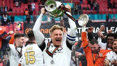 Byron Webster holds the National League promotion final winners' trophy aloft cheering, while teammates around him celebrate 