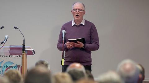 A man singing from a hymn book. He has grey, receded hair and glasses and is wearing a purple jumper with a light coloured shirt underneath. He is standing in front of a microphone