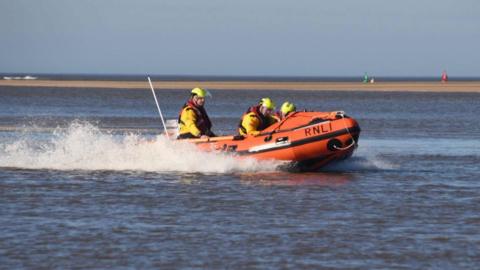 Three people onboard a small orange lifeboat which is travelling at speed across the water.