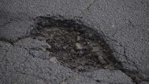 A close‑up view of a pothole in a road surface. It shows cracked edges and exposed loose stones within the damaged tarmac.