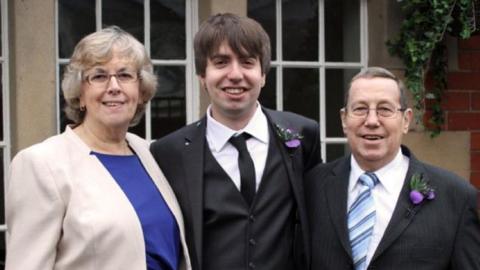 A man wearing his wedding suit, standing with his mother and father either side, in front of the windows of a building.  