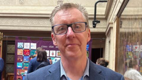 Stephen Thompson looking at the camera. He is wearing a blue blazer, light blue shirt and a blue lanyard around his neck. He is wearing glasses.