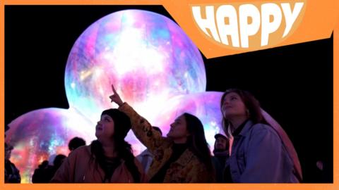 People at a light festival and the Happy News logo