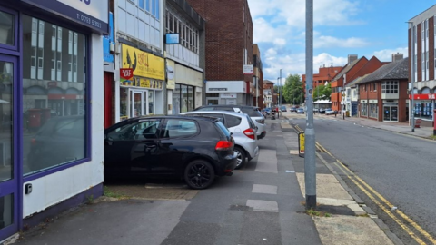 Image shows cars parked on a pavement with the backs of the cars protruding into the pavement. The road next to the pavement is clear and the cars are parked facing shops.