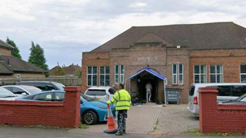 A Google streetview image of the mosque building from outside - with cars parked in front of the building. A person in a yellow high-vis jacket is standing at the entrance to the car park, holding an orange traffic cone.