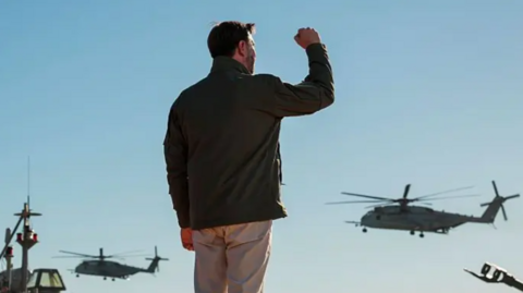 JD Vance with his back to the camera raises his right arm in celebration as two helicopters fly overhead