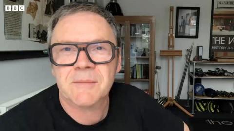 Brian Cannon, who is wearing a black t-shirt and black, square-rimmed glasses, sits in his studio on a Zoom call to the BBC