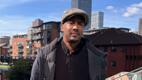 Paul Bridgewater is wearing a grey jacket and tweed cap standing outside the Mailbox in Birmingham with city buildings in the background